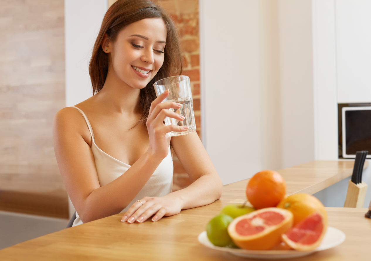 Happy Young Woman Holding Drinking Water Glass while there is Some Fruits in Front of her