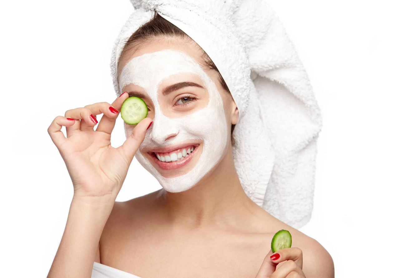 Smiling Young Girl with Facial Mask on her Face is Holding a Slice of Cucumber in Front of her Eye