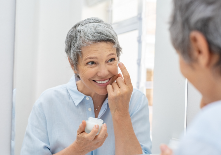 Smiling Middle-Aged Woman Standing in front of the Mirror and Applying Cream on Face